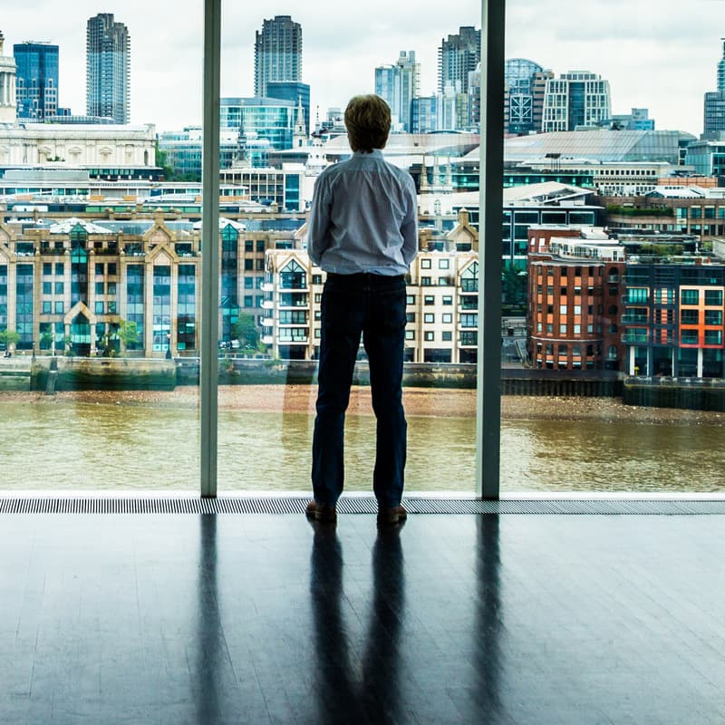 man looking out a window at the people crossing the river by walking over a bridge