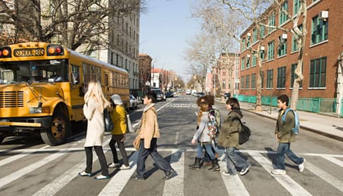 school kids in crosswalk with teacher. School bus in the background. Urban environment.